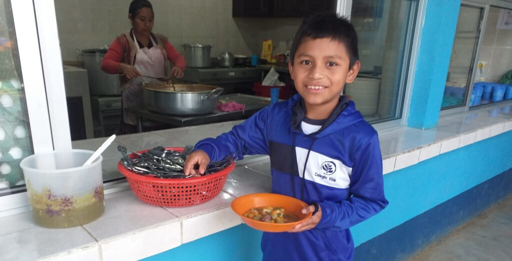 AxelReceivingSchoolMeal-WEB Boy in blue school uniform holding plate of food at cafeteria window. Woman cooking in the background.