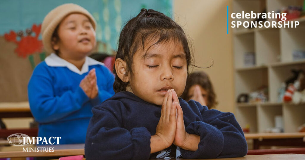 A Guatemalan girl with her eyes closed, hands folded and head bowed praying at her desk. Behind is another child in a blue Vida school sweater and a tan hat also praying