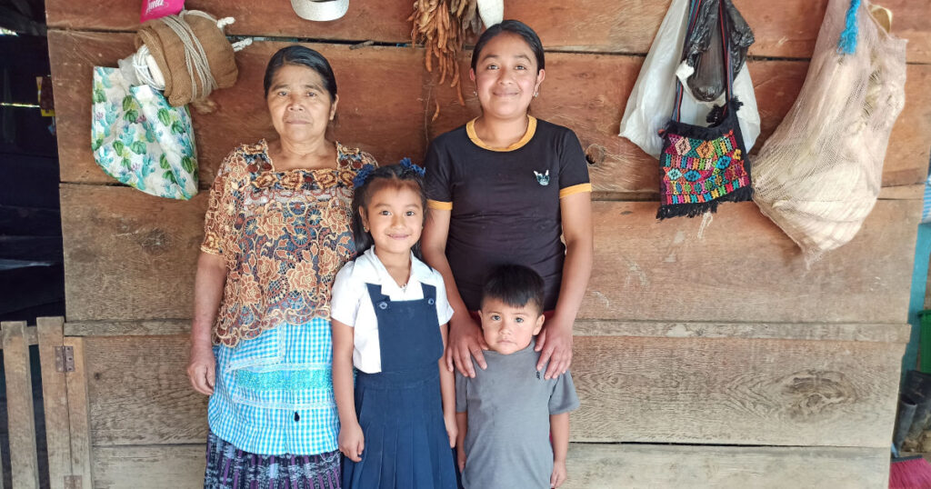 Guatemalan woman in t-shirt standing behind two children, a young girl in school uniform and an even younger boy. Next to them is an older woman in traditional guatemalan dress